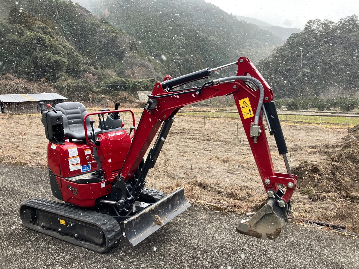 A small excavator standing in a bare field in falling snow, with forested mountains behind.